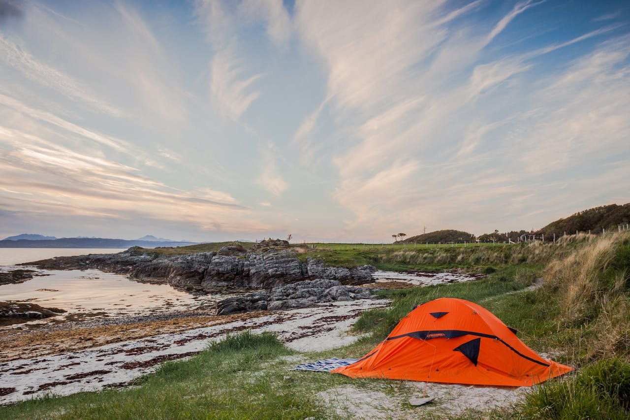 Orange tent set by the serene coast in Highland, Scotland during calm summer evening.
