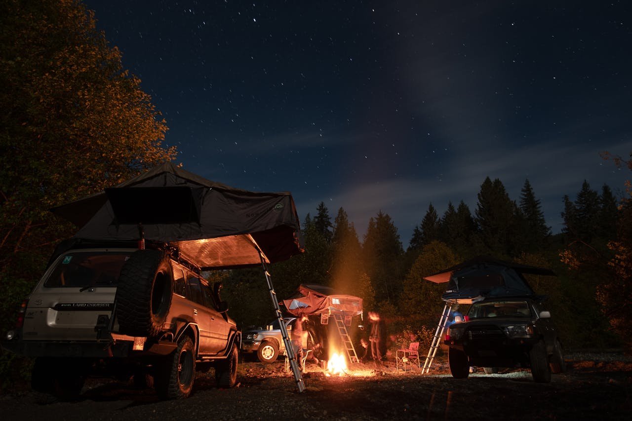Camping scene with cars and tents under a starry night sky, featuring a cozy bonfire in the forest.